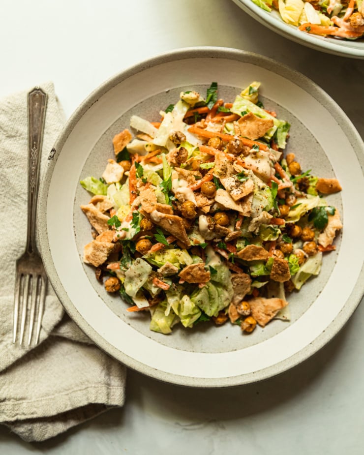 An overhead shot shows an individual serving of a hummus crunch salad, featuring: iceberg lettuce, matchstick carrots, parsley, za'atar chickpeas, crushed up pita chips, and a creamy hummus-based dressing. Salad tongs are nearby the bowl.