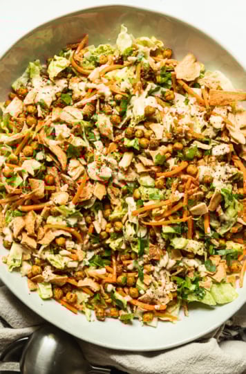 An overhead shot shows a large serving bowl filled with hummus crunch salad, featuring: iceberg lettuce, matchstick carrots, parsley, za'atar chickpeas, crushed up pita chips, and a creamy hummus-based dressing. Salad tongs are nearby the bowl.