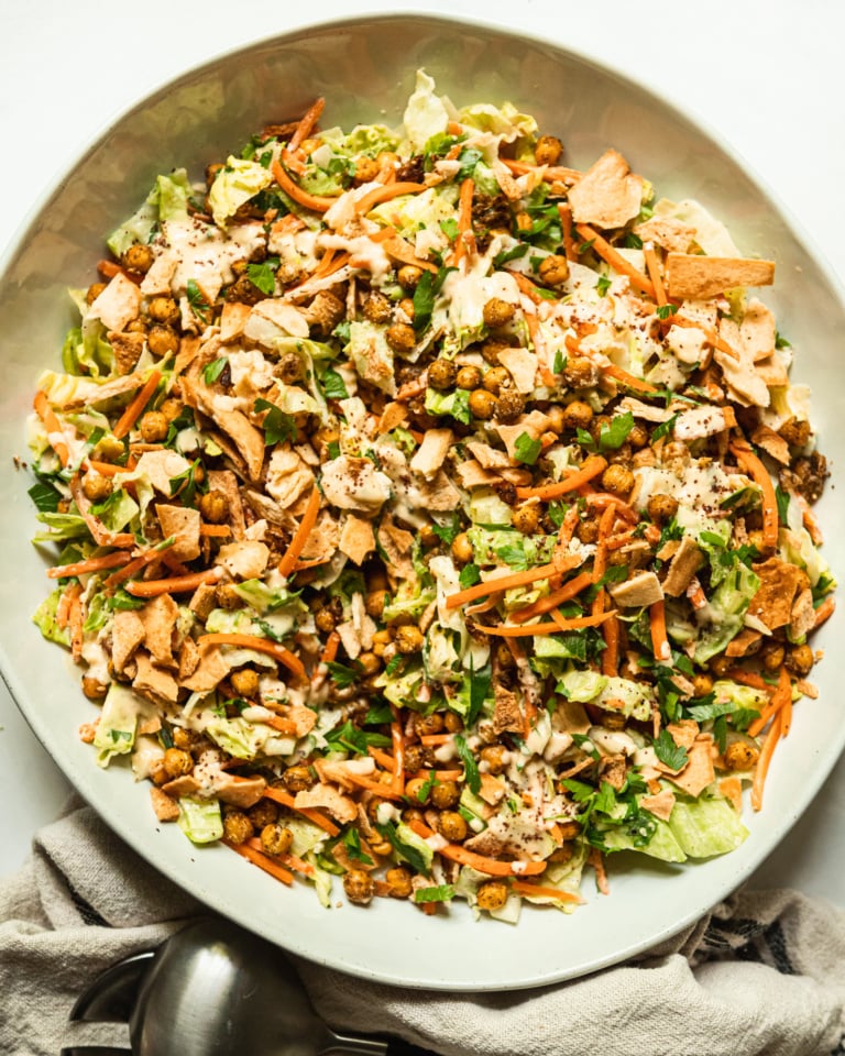 An overhead shot shows a large serving bowl filled with hummus crunch salad, featuring: iceberg lettuce, matchstick carrots, parsley, za'atar chickpeas, crushed up pita chips, and a creamy hummus-based dressing. Salad tongs are nearby the bowl.