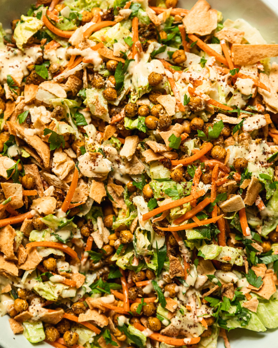 An up close, overhead shot shows a large serving bowl filled with hummus crunch salad, featuring: iceberg lettuce, matchstick carrots, parsley, za'atar chickpeas, crushed up pita chips, and a creamy hummus-based dressing.