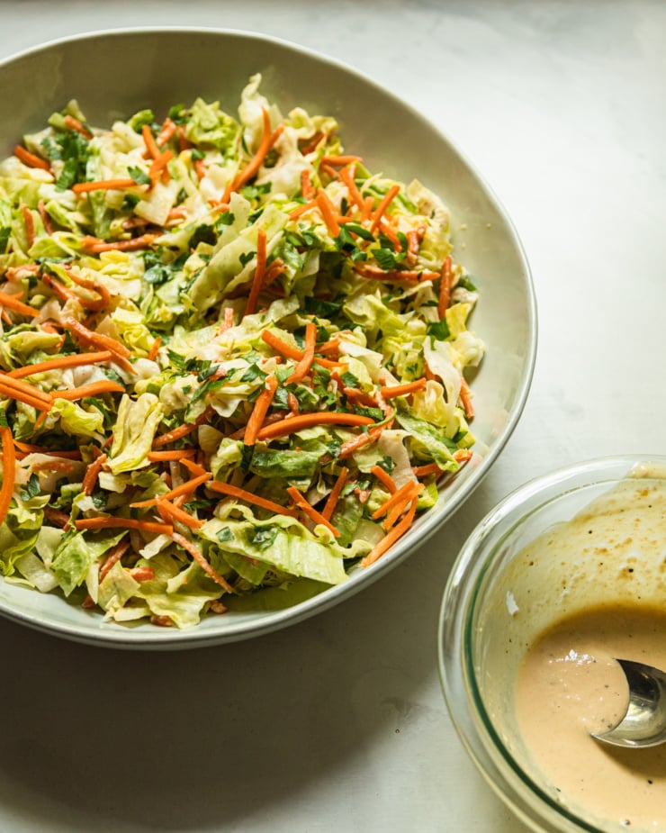 A 34 angle shot shows a mized green salad with carrot matchsticks and a bowl of creamy dressing nearby.