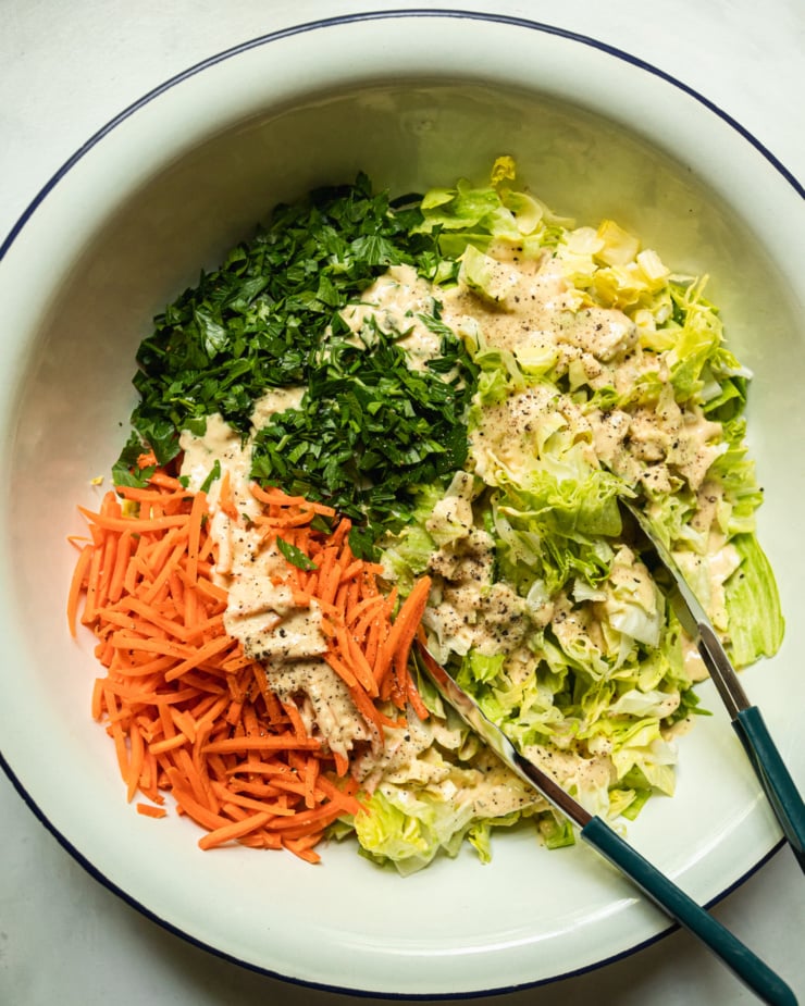 An overhead shot shows a large salad bowl filled with chopped iceberg lettuce, matchstick carrots, and chopped parsley. The vegetables are drizzled with a creamy dressing and tongs are sticking out of the bowl.