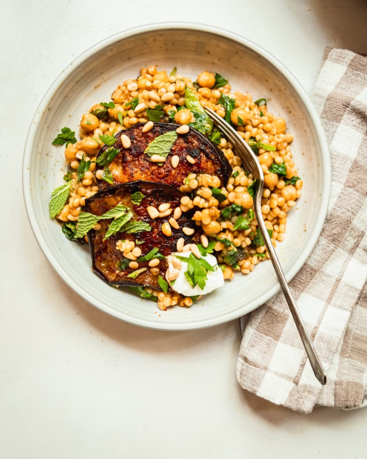 An overhead shot shows an individual serving of pearl couscous and chickpeas with slices of harissa eggplant on top. The dish includes chopped spinach and herbs as well.