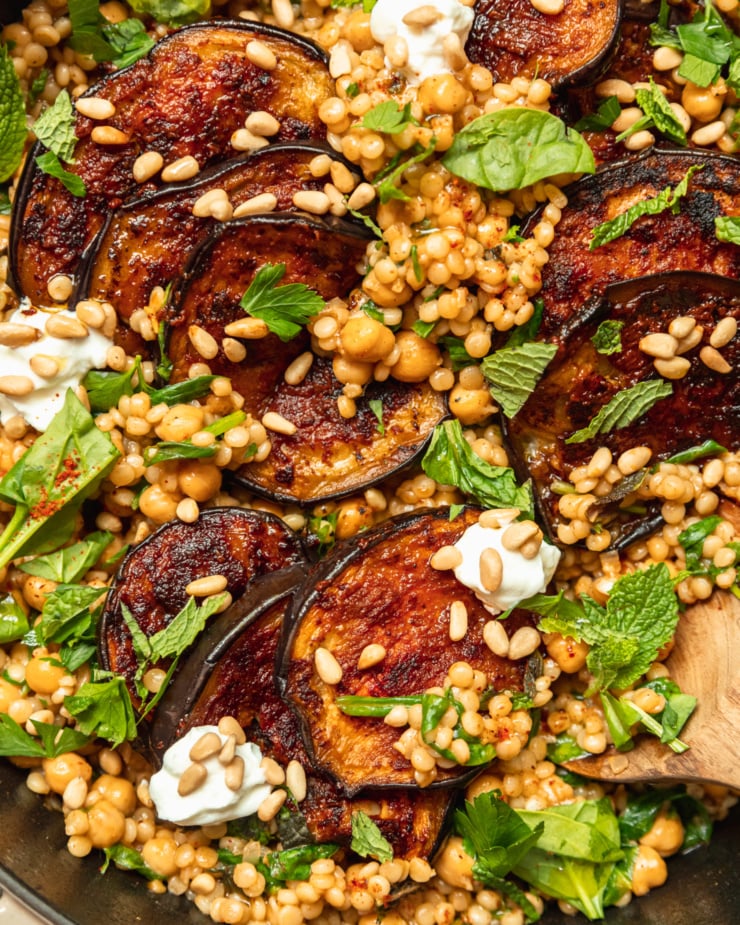 An up close, overhead shot of one-pan harissa eggplant with pearl couscous, chickpeas, spinach, herbs, pine nuts, and dollops of vegan yogurt.