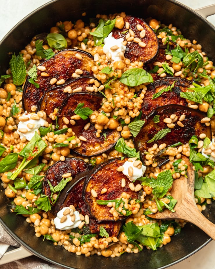 An up close, overhead shot of one-pan harissa eggplant with pearl couscous, chickpeas, spinach, herbs, pine nuts, and dollops of vegan yogurt.