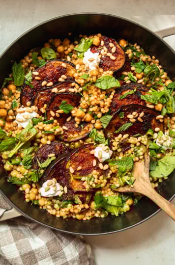 An overhead shot of one-pan harissa eggplant with pearl couscous, chickpeas, spinach, herbs, pine nuts, and dollops of vegan yogurt. The dish is shown in a cast iron braiser-style pot and a wooden spoon is sticking out.