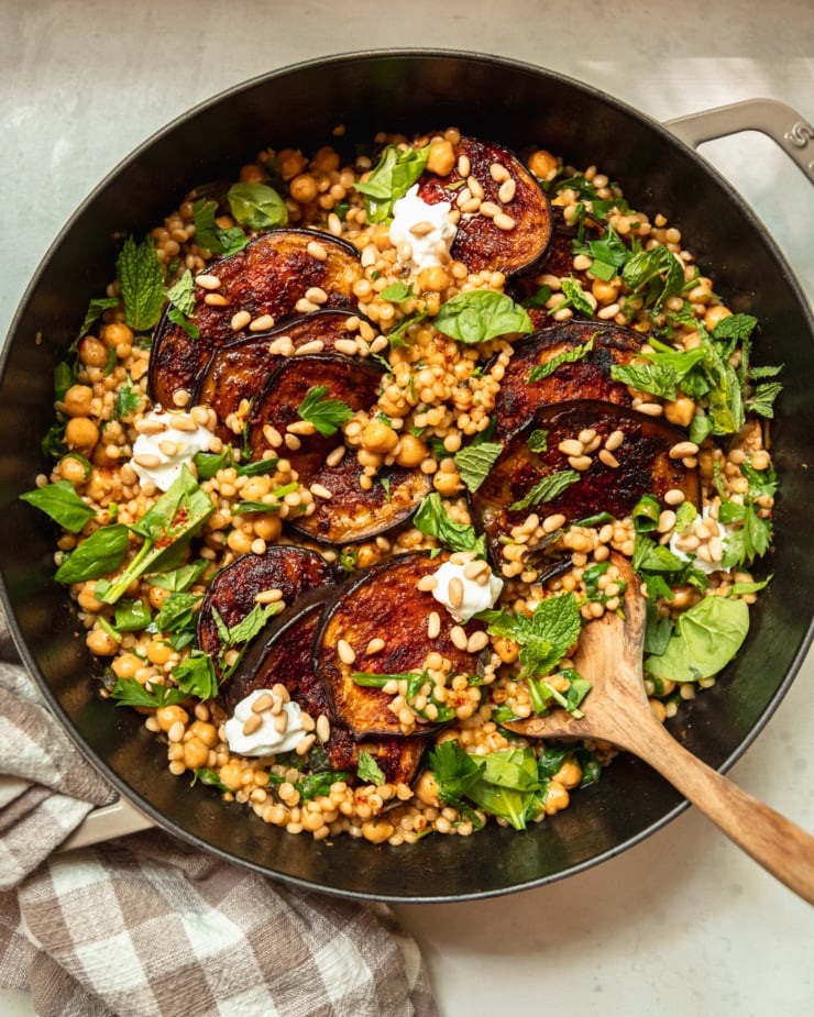 An overhead shot of one-pan harissa eggplant with pearl couscous, chickpeas, spinach, herbs, pine nuts, and dollops of vegan yogurt. The dish is shown in a cast iron braiser-style pot and a wooden spoon is sticking out.