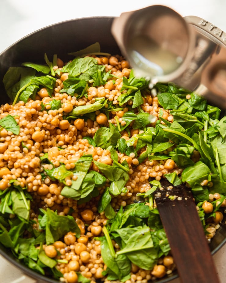 A 3/4 angle shot shows pearl couscous, chickpeas, and spinach together in a pot. Lemon juice is being added to the mixture from a measuring cup.