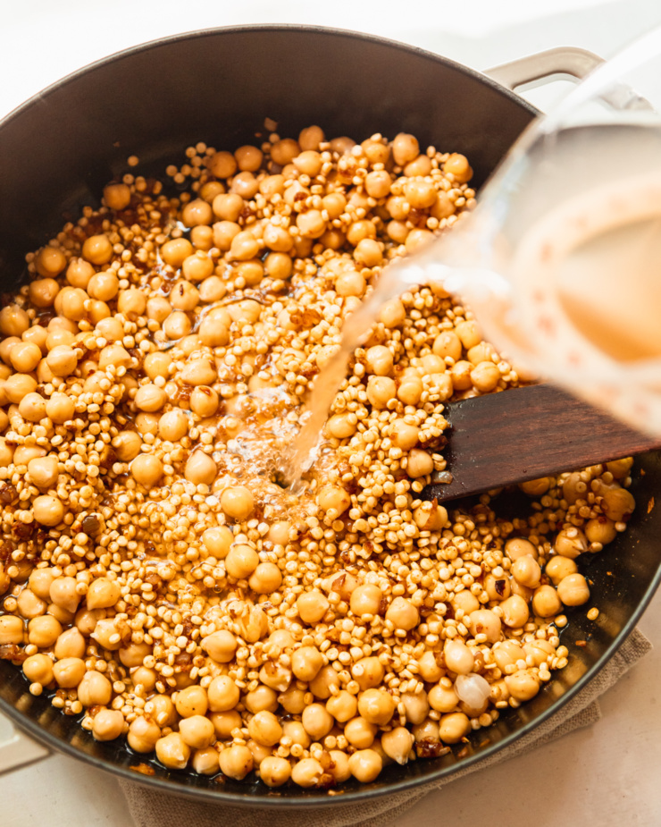 An overhead shot shows vegetable stock being added to a pot of chickpeas and pearl couscous.