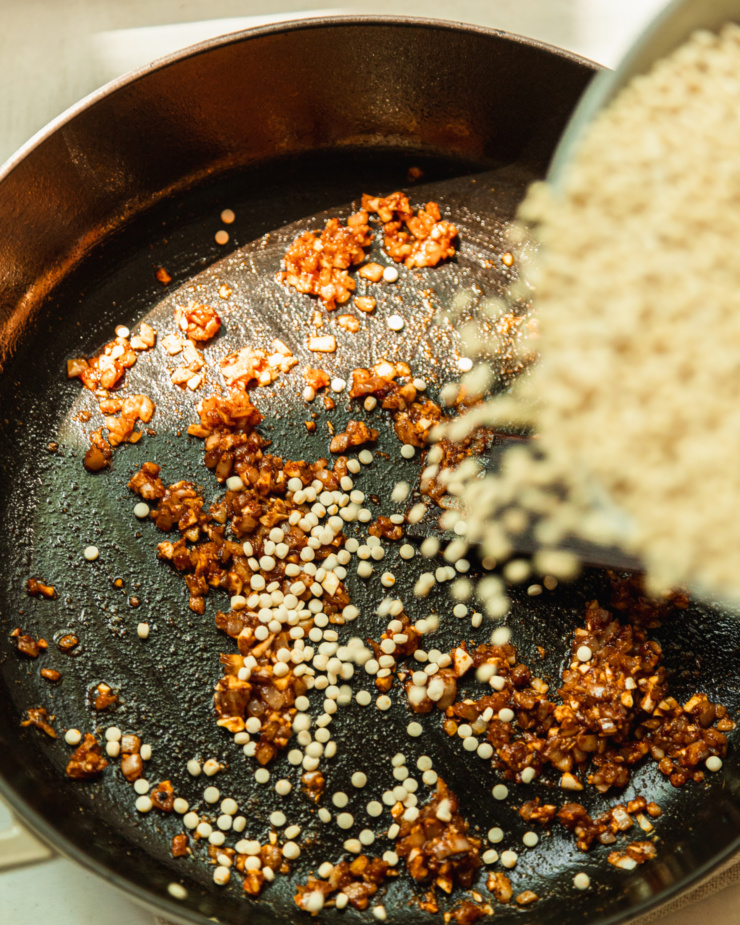 An overhead shot shows pearl couscous being added to a hot pan with sautéed shallots, garlic, and spices.