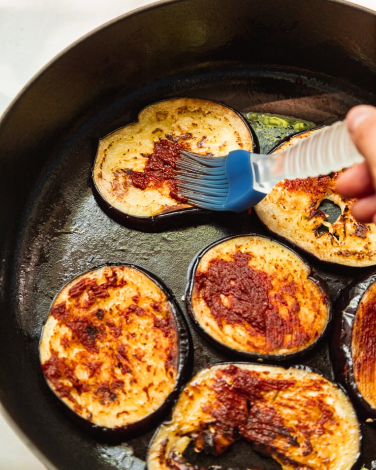An overhead shot shows a hand using a brush to apply harissa paste to sautéed slices of eggplant in a braiser-style pot.