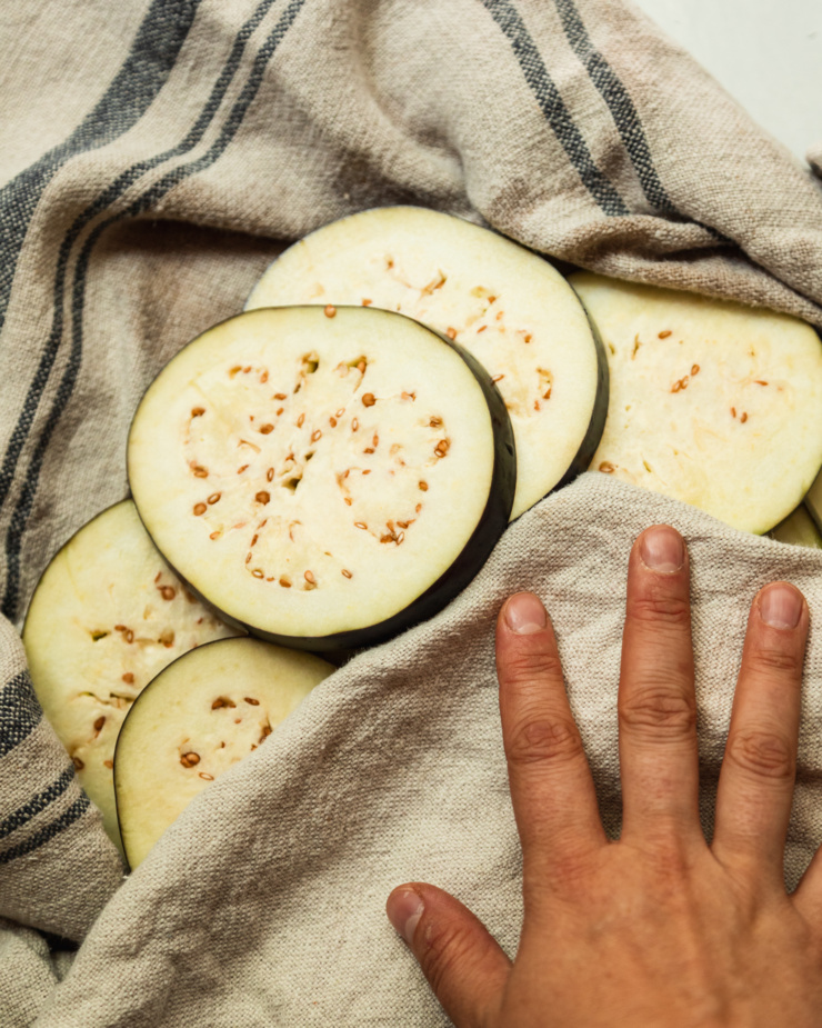 An overhead shot shows slices of eggplant being pat dry with a kitchen towel.