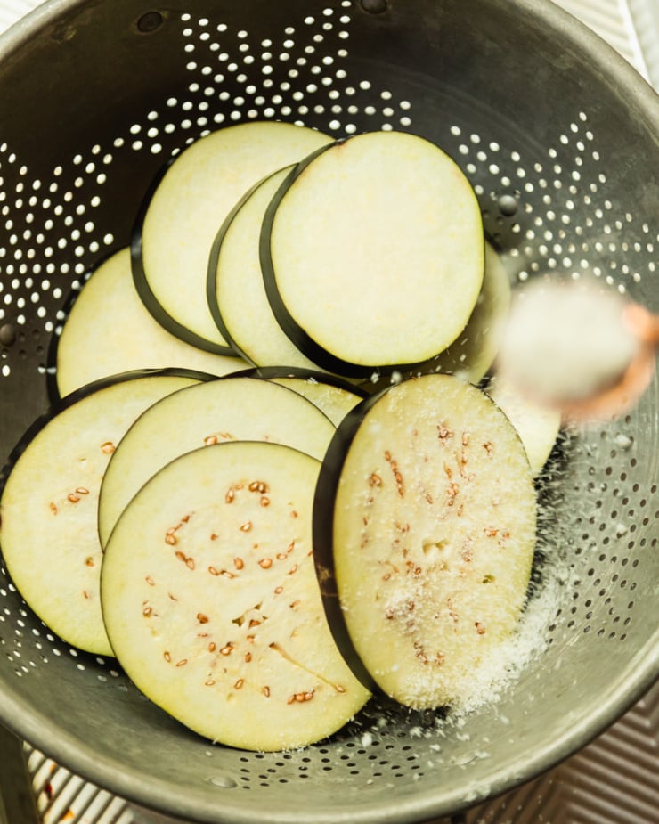 An overhead shot shows a spoonful of salt being added to slices of eggplant in a colander.