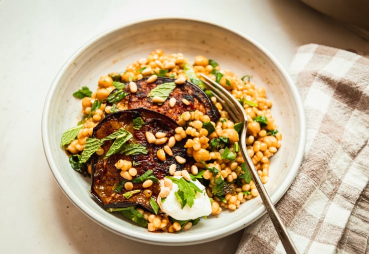 A 3/4 angle image shows an individual serving of pearl couscous and chickpeas with slices of harissa eggplant on top. The dish includes chopped spinach and herbs as well.