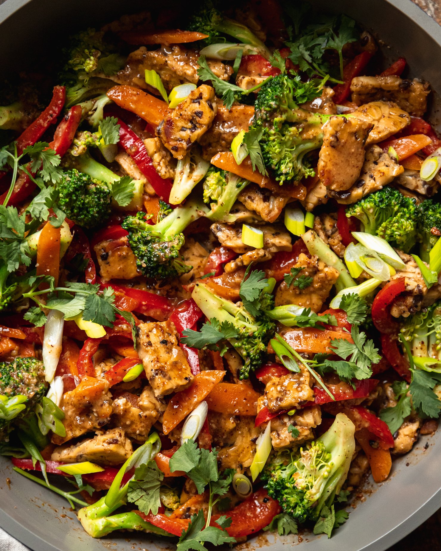 An up close, overhead shot of an almond butter tempeh stir fry with vegetables in a nonstick frying pan. The stir fry is topped with sliced green onions and chopped cilantro.