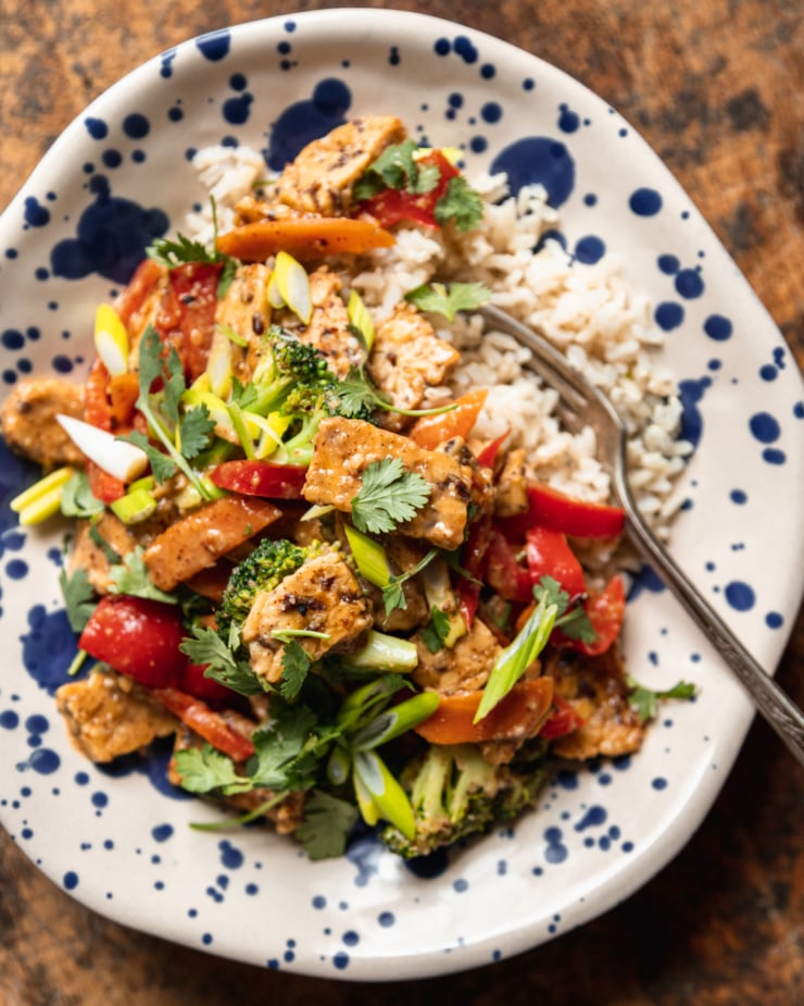 An overhead shot of a serving of almond butter tempeh stir fry with vegetables over a pile of cooked brown rice. The single serving is in an off-white bowl with free-painted blue dots all over it.