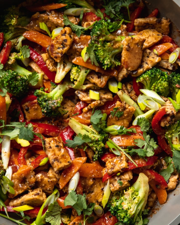 An up close, overhead shot of an almond butter tempeh stir fry with vegetables in a nonstick frying pan. The stir fry is topped with sliced green onions and chopped cilantro.