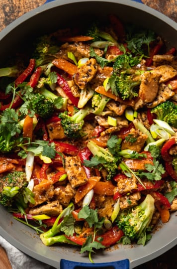 An up close, overhead shot of an almond butter tempeh stir fry with vegetables in a nonstick frying pan. The stir fry is topped with sliced green onions and chopped cilantro.