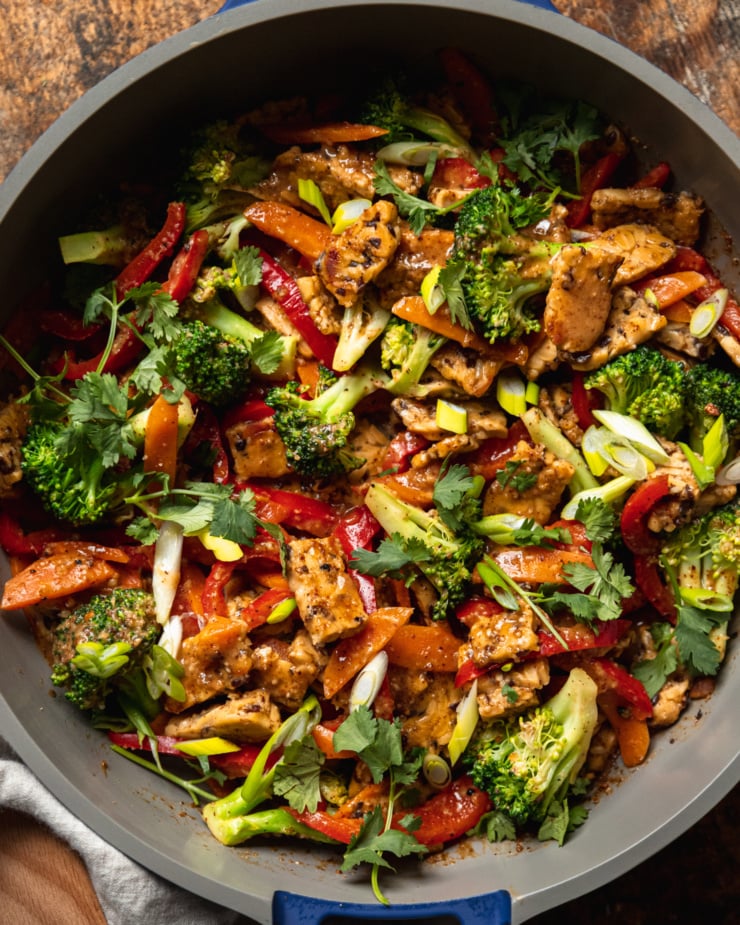 An up close, overhead shot of an almond butter tempeh stir fry with vegetables in a nonstick frying pan. The stir fry is topped with sliced green onions and chopped cilantro.