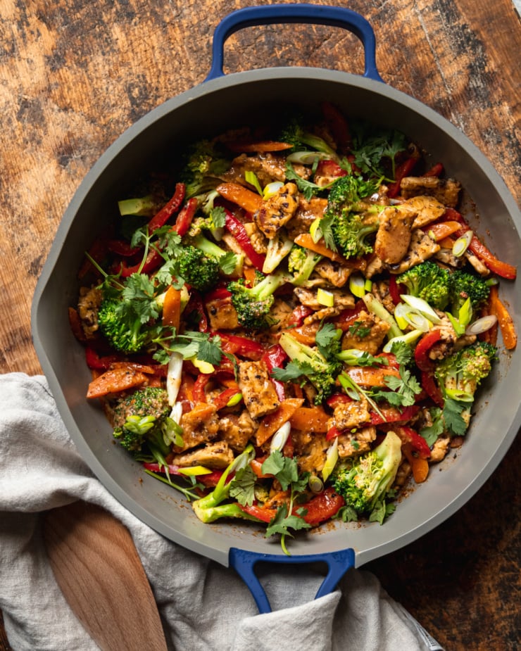 An overhead shot of an almond butter tempeh stir fry with vegetables in a nonstick frying pan. The stir fry is topped with sliced green onions and chopped cilantro.