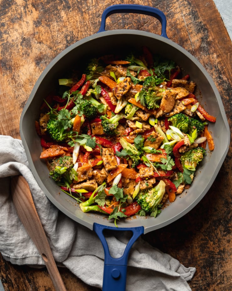 An up close, overhead shot of an almond butter tempeh stir fry with vegetables in a nonstick frying pan. The stir fry is topped with sliced green onions and chopped cilantro.