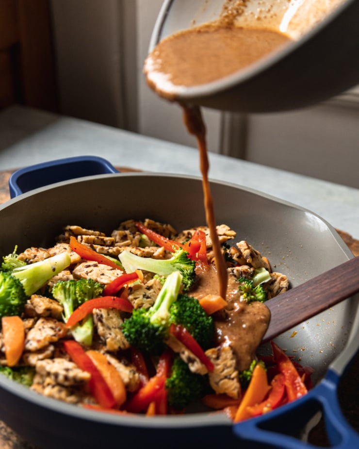 A 3/4 angle shot shows almond butter sauce being poured into a frying pan filled with stir fried vegetables and pieces of tempeh.