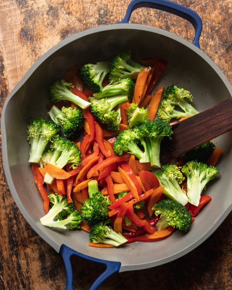 An overhead shot shows broccoli florets, red pepper strips, and sliced carrots being stir fried in a frying pan.