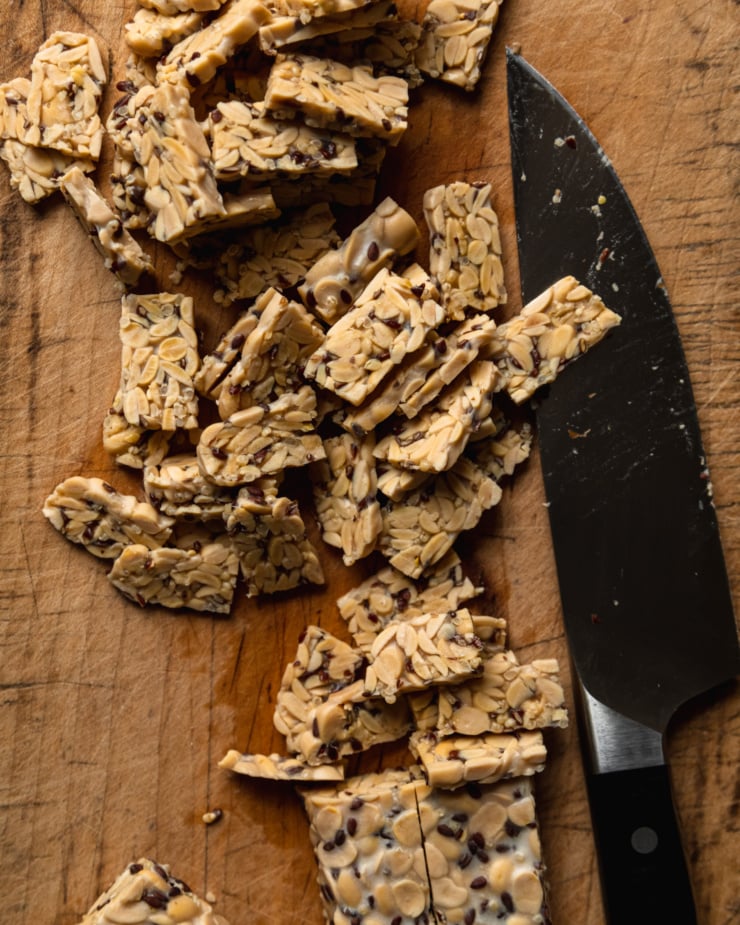 An up close, overhead shot shows pieces of tempeh being thinly sliced on a wooden cutting board with a chef's knife.