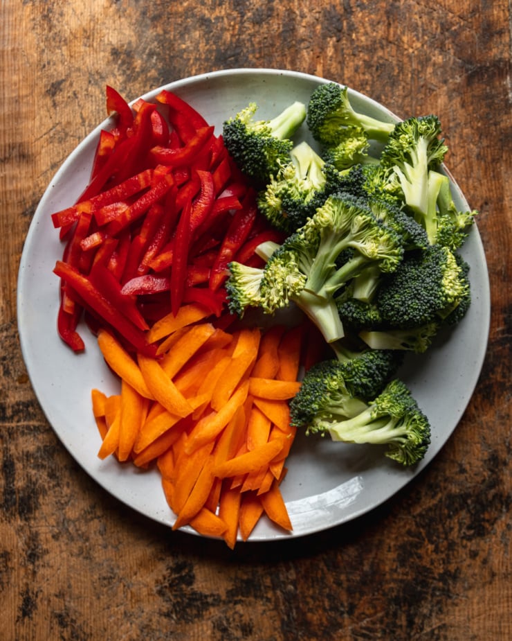 An overhead shot shows sliced red bell peppers, sliced carrots, and broccoli florets prepped and ready on a plate.