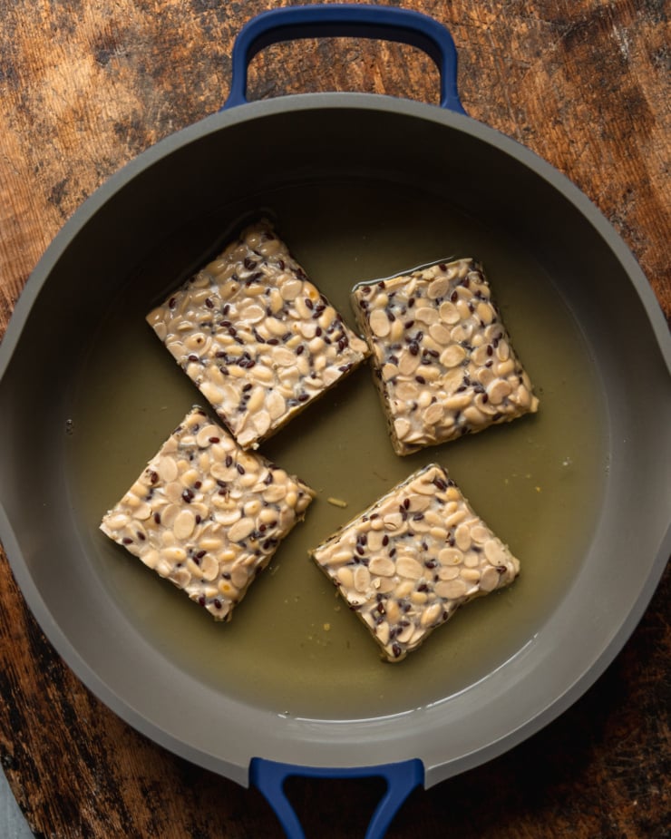 An overhead shot shows 4 pieces of tempeh being steamed/boiled in a frying pan filled with water.