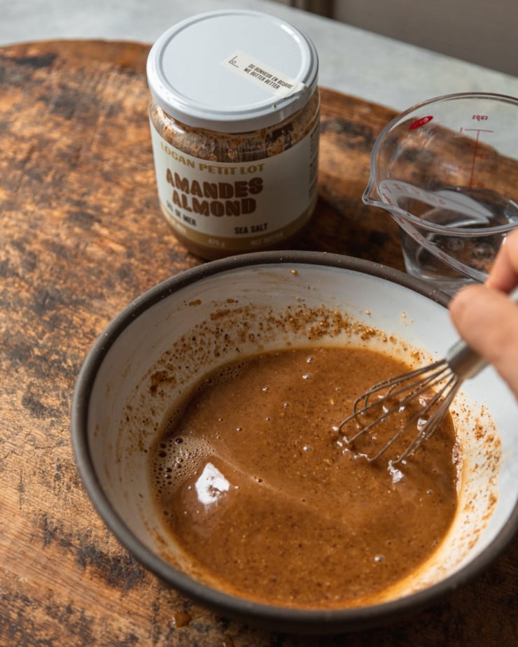 A 3/4 angle shot shows a hand using a whisk to stir together some almond butter sauce. The jar of almond butter is in the background.