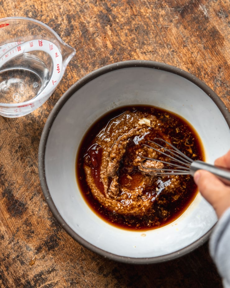 An overhead shot shows a hand using a whisk to bring together ingredients for an almond butter sauce in a medium-sized ceramic bowl.