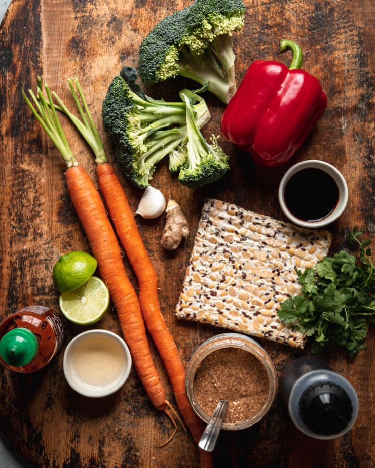 An overhead shot shows ingredients used in an almond butter tempeh stir fry with vegetables. All ingredients are set against a worn wood background.