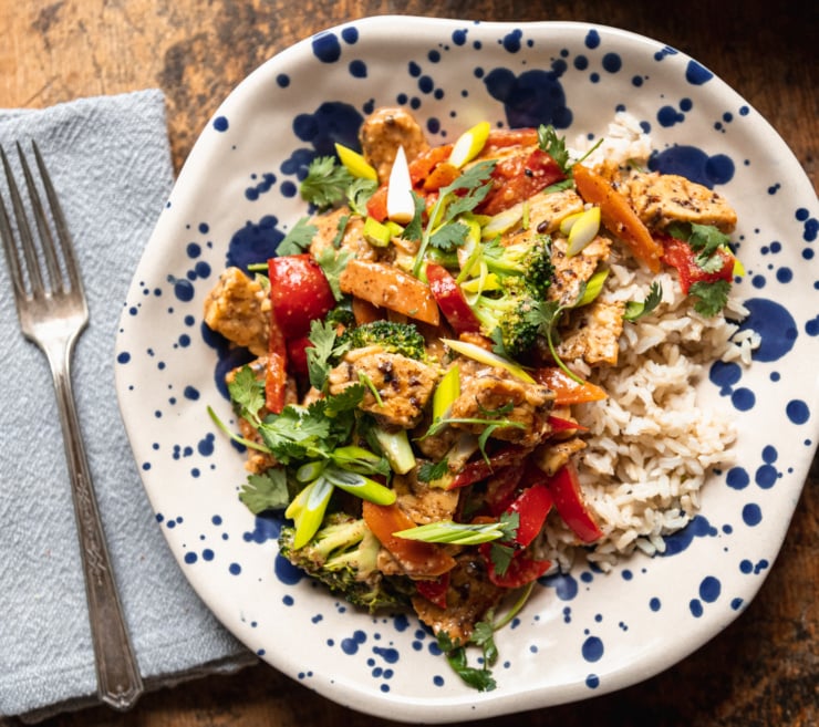 An overhead shot of a serving of almond butter tempeh stir fry with vegetables over a pile of cooked brown rice. The single serving is in an off-white bowl with free-painted blue dots all over it.