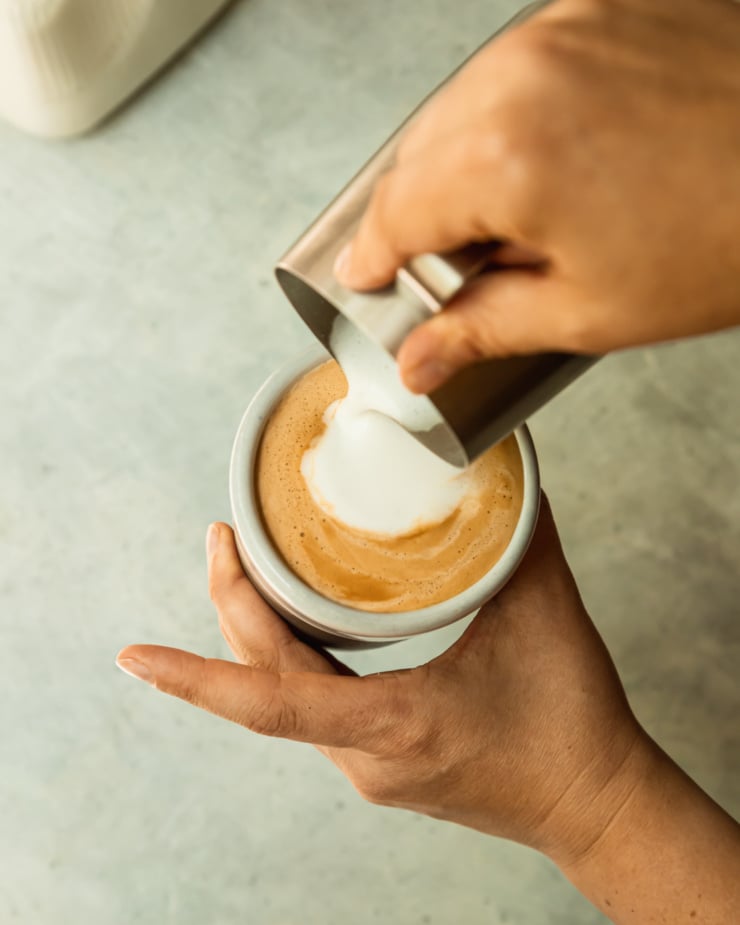 An overhead shots a hand pouring frothy milk into a cup of espresso.
