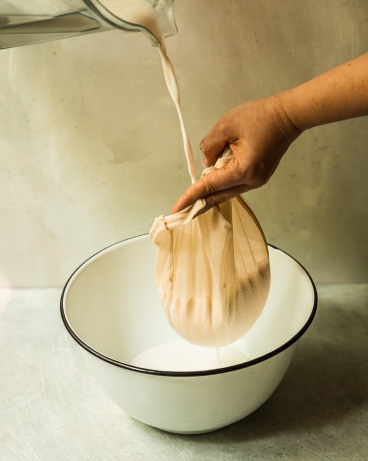 A 3/4 angle shot shows nut milk being poured into a nut milk bag that is positioned over a large enamelware bowl.