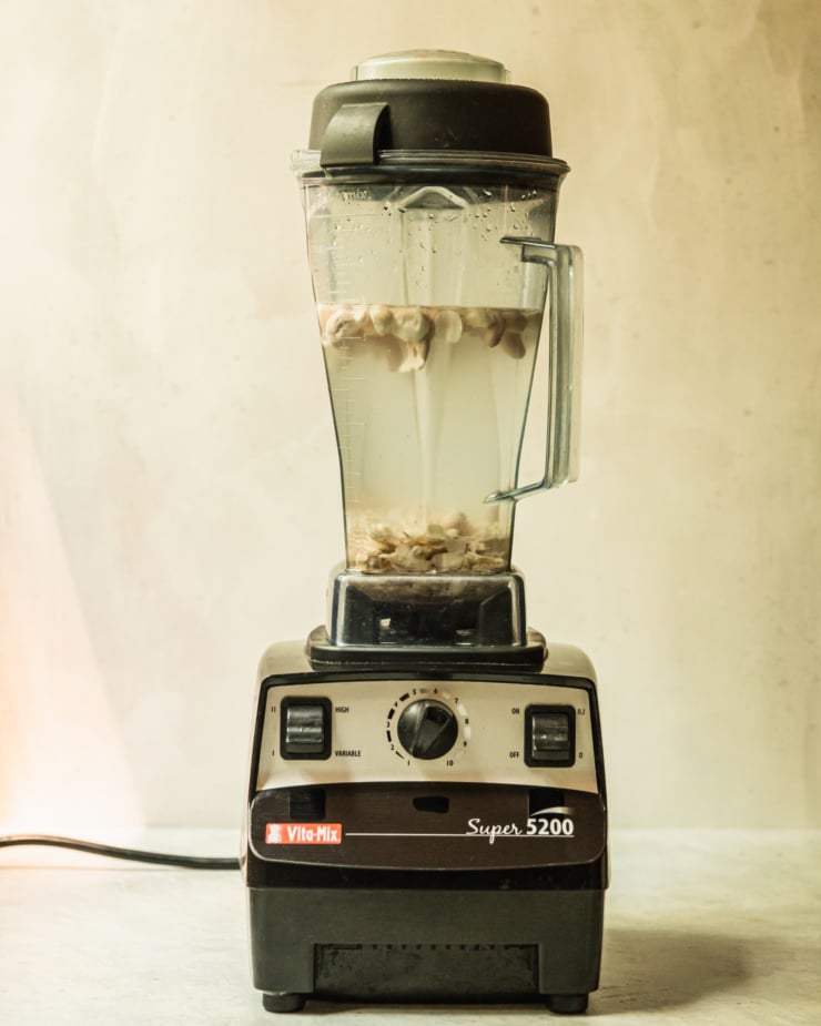 A head-on shot shows a Vitamix blender with the pitcher containing water and soaked cashews, macadamia nuts, and sliced almonds.