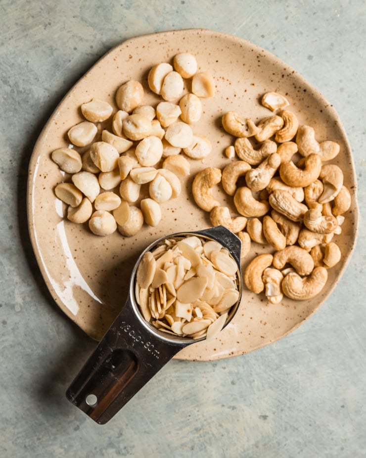An overhead shot shows three types of raw nuts on a plate: macadamia, cashews, and sliced almonds.