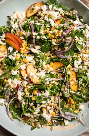 An overhead shot of a fresh summer salad featuring: cooked corn, sliced peaches, arugula, pumpkin seeds, spicy cashew dressing, and vegan feta.