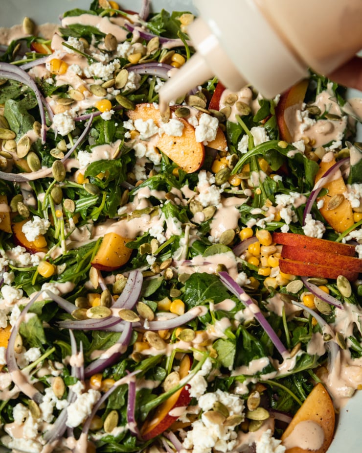 An up close, overhead shot of a fresh summer salad featuring: cooked corn, sliced peaches, arugula, pumpkin seeds, spicy cashew dressing, and vegan feta. The dressing is being applied with a squeeze bottle.
