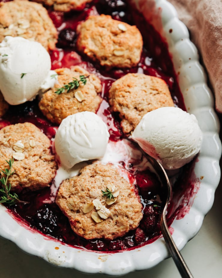 A 3/4 angle shot shows a baking dish filled with vegan berry cobbler and topped with 3 scoops of melt-y vegan vanilla ice cream.