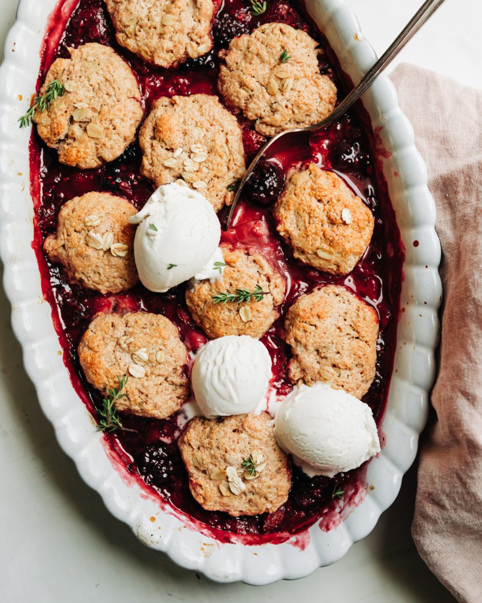 An overhead shot shows a baked vegan berry cobbler topped with 3 scoops of vanilla vegan ice cream and little time leaves. A spoon is sticking out of the baking dish.