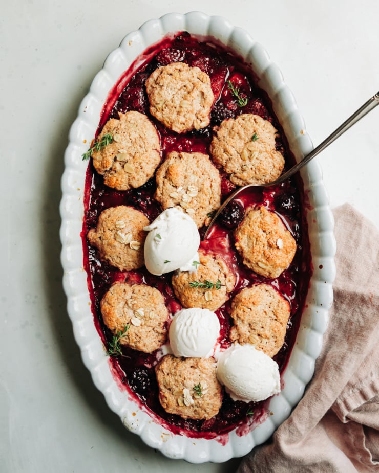 An overhead shot shows a baked vegan berry cobbler topped with 3 scoops of vanilla vegan ice cream and little time leaves. A spoon is sticking out of the oval, scallop-edged baking dish.