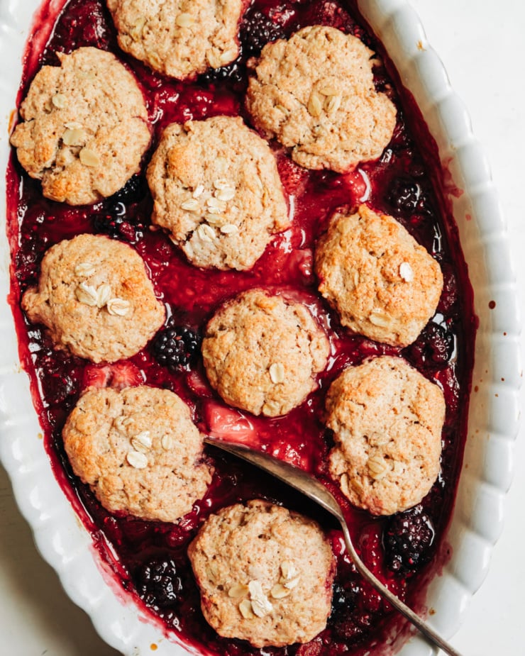 An overhead shot shows a baked vegan berry cobbler with oat-y biscuit topping.