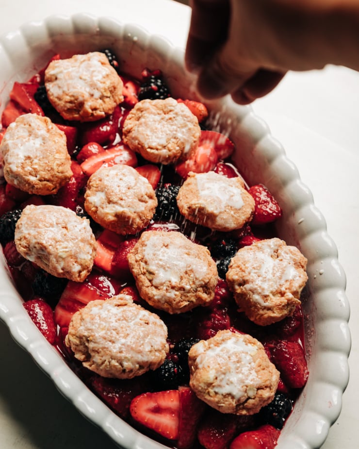 A 3/4 angle shot shows a hand sprinkling sugar on top of oat-y biscuits that top a fruit cobbler.