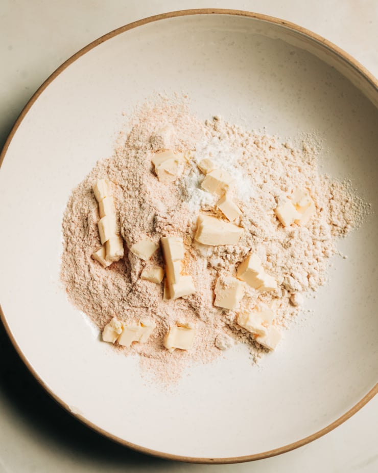 An overhead shot shows spelt flour, baking powder, salt, and diced vegan butter in a mixing bowl.