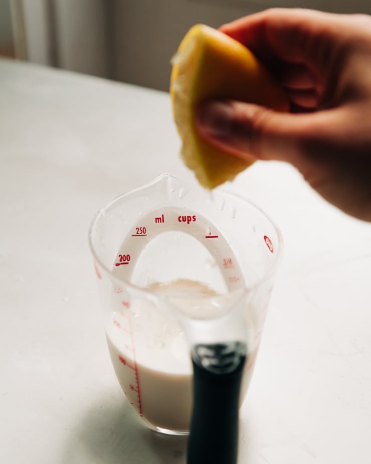 A head-on shot shows a hand squeezing a lemon into a measuring cup of soy milk.