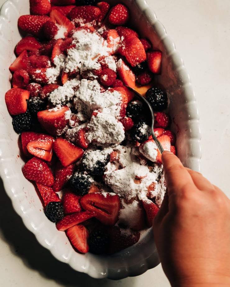 An overhead shot shows a hand using a spoon to stir some arrowroot and sugar into some chopped berries.