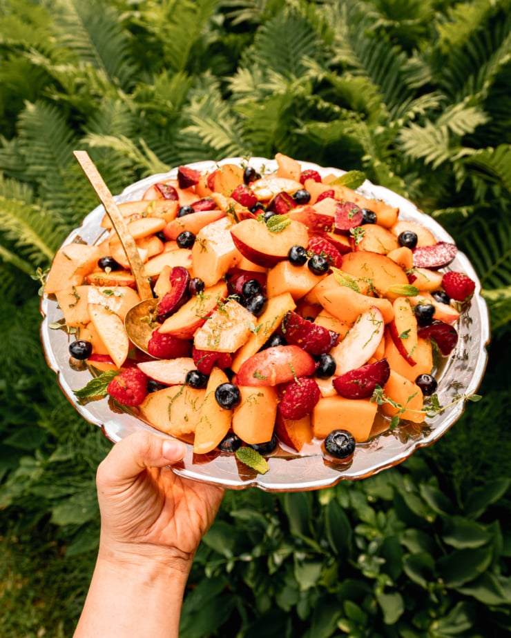 A 3/4 angle shots an arm extended out, holding a platter of fruit with fresh mint on top. Wild ferns are seen in the background.