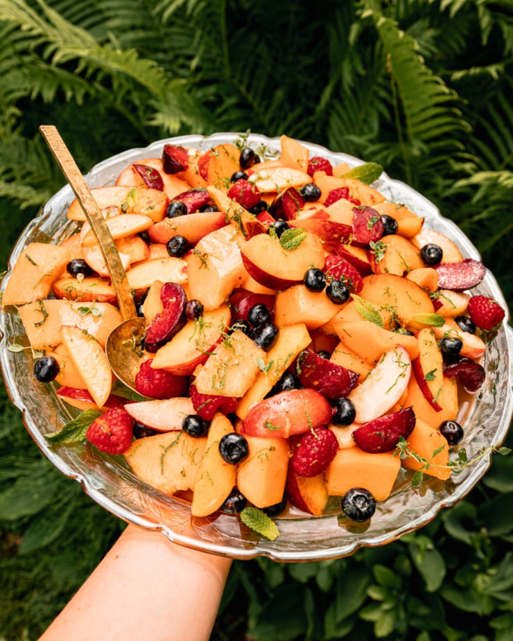 A 3/4 angle shots an arm extended out, holding a platter of fruit with fresh mint on top. Wild ferns are seen in the background.